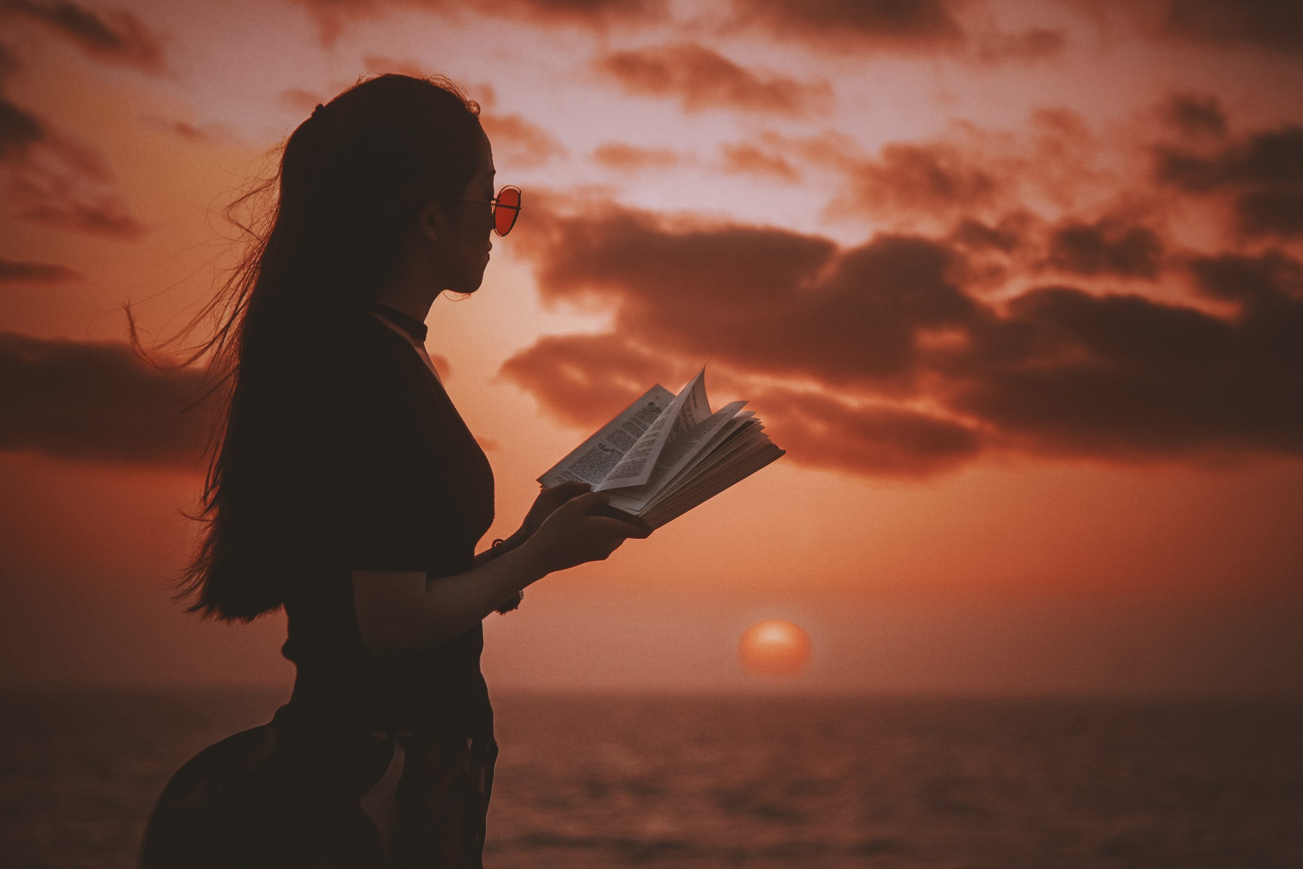 Eine Frau liest ein Buch und steht am See. Im Hintergrund geht die Sonne runter.