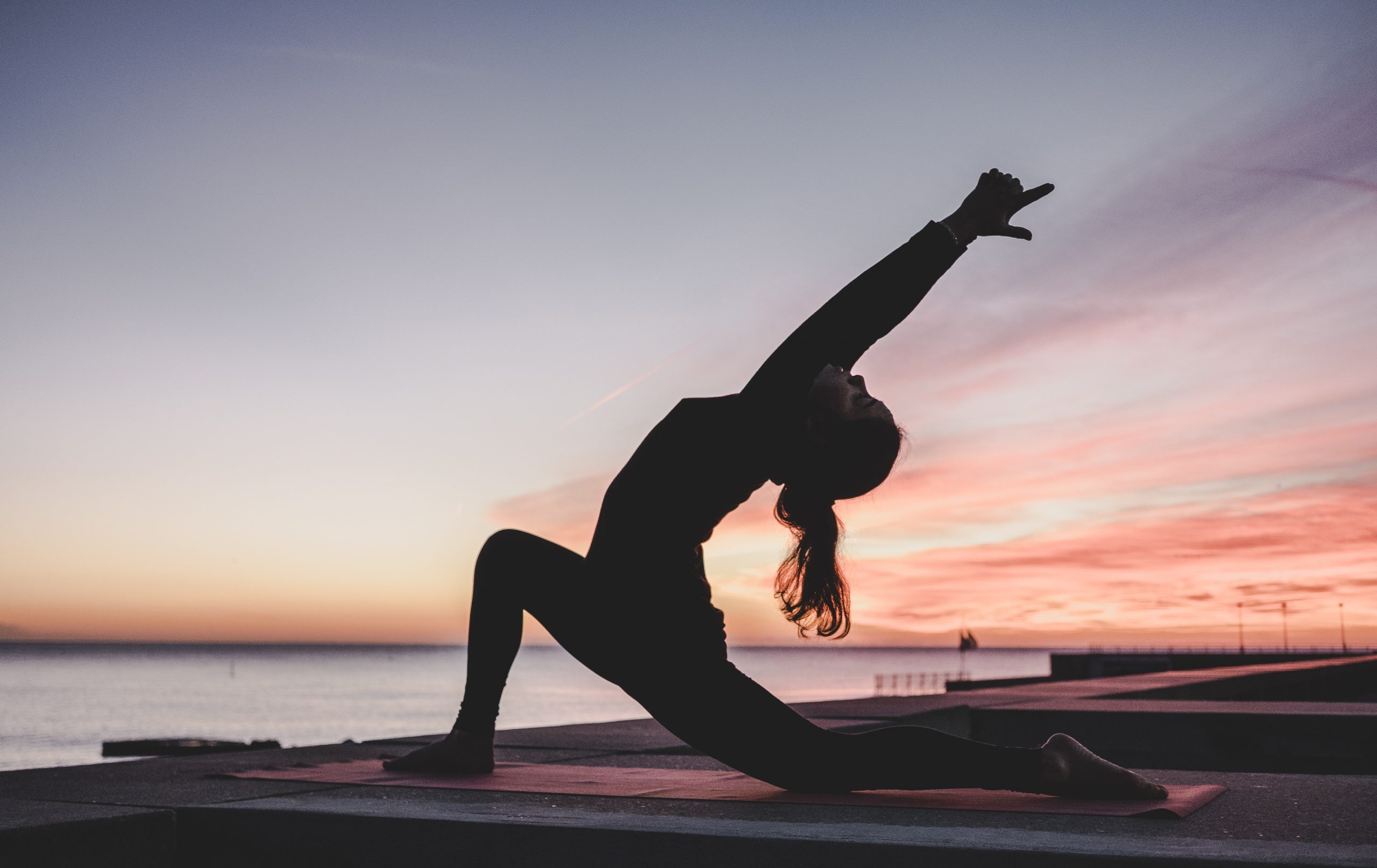 Eine Frau macht Yoga Übungen am See. Im Hintergrund leuchtet der Himmel im abendrot des Sonnenuntergangs.