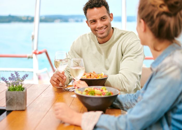 Ein junges Paar sitzt an einem Tisch auf einer Fähre der BSB. Sie halten beide ein Glas Wein in der Hand. Vor ihnen steht ein Teller mit frischem Essen. Der Mann lächelt seine Frau an. Im Hintergrund ist der Bodensee zu sehen.