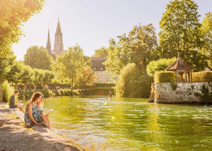 Eine Frau sitzt am Gewässer im Stadtgarten Konstanz und liest ein Buch. Im Hintergrund scheint die Sonne und das Konstanzer Münster ist zu sehen.