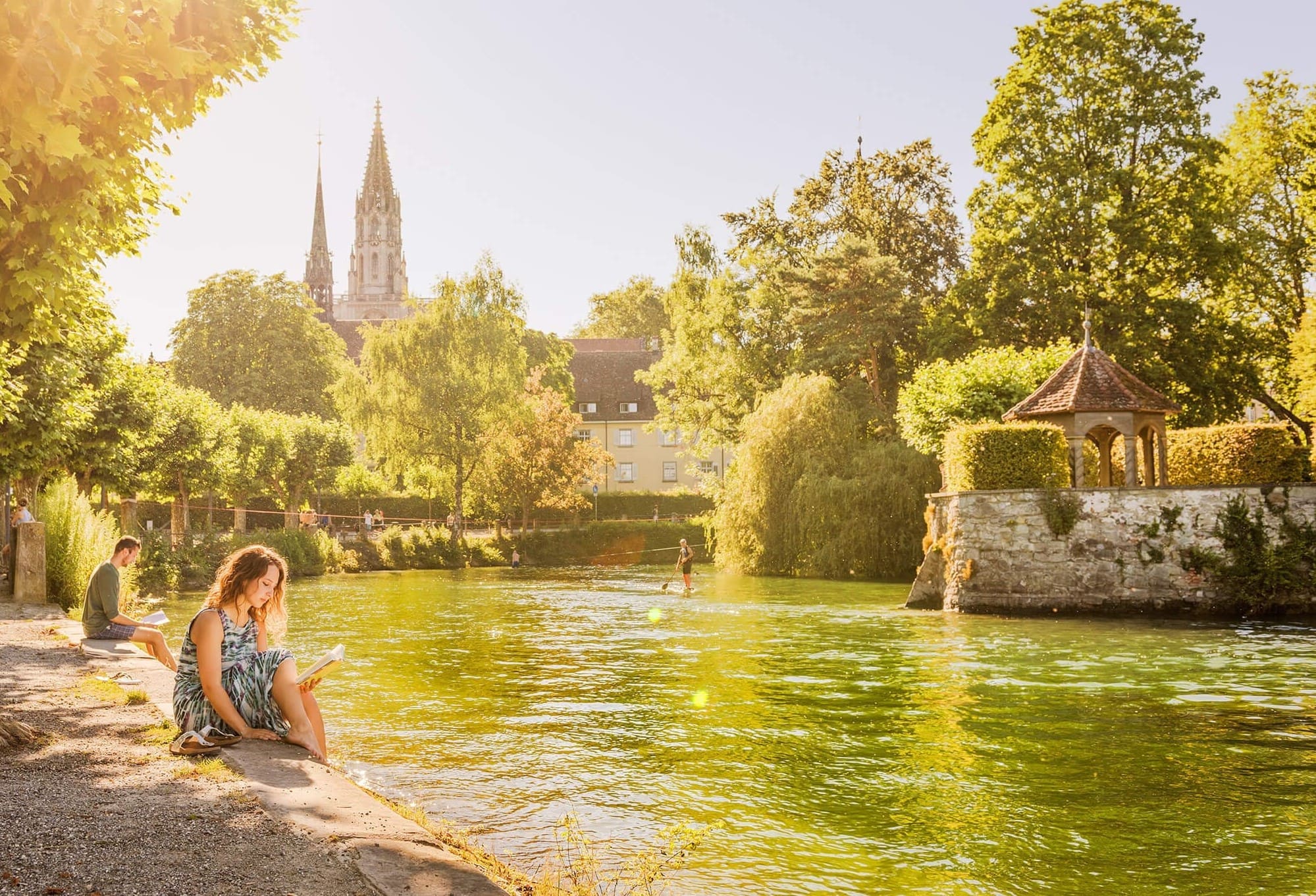 Eine Frau sitzt am Gewässer im Stadtgarten Konstanz und liest ein Buch. Im Hintergrund scheint die Sonne und das Konstanzer Münster ist zu sehen.